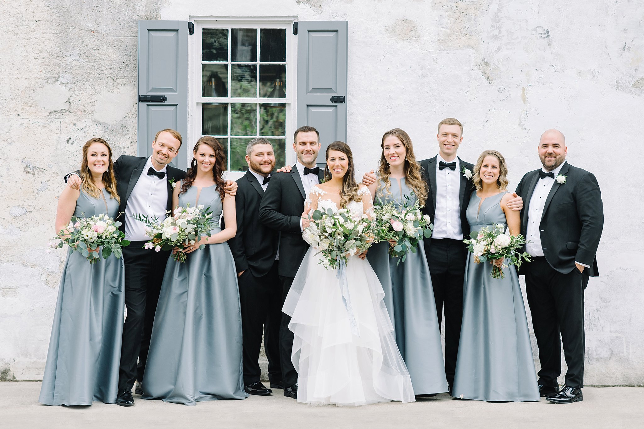 Bride and groom with their bridal party in downtown Charleston. Black tie wedding. Wedding gown with lace sleeves. Blue floor length bridesmaid gowns